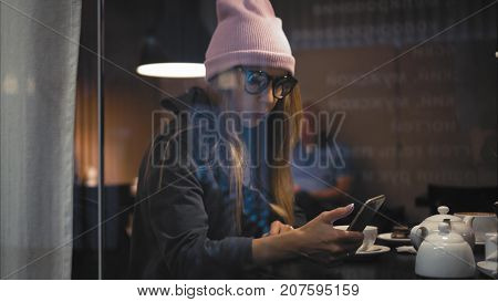 A stylish hipster female in glasses using smart watch and using app on a smartphone in a cafe. View through the window