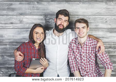 Three smiling european friends with tablet standing on wooden wall background. Friendship concept