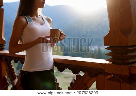 Slim caucasian woman holds cup of tea in her hands at mountain resort. Sports girl with hot coffee mug at wooden balcony of country house. Forest and mountains on background Dietary concept.