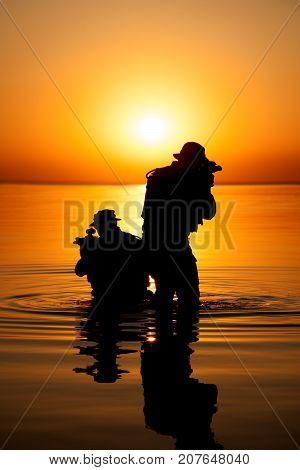 Army soldiers with rifles orange sunset silhouette in action during raid crossing river in the water