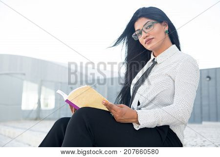 Confident female entrepreneur reading business book to achieve success. Serious intelligent Hispanic lady resting with book outdoors. Self-education concept
