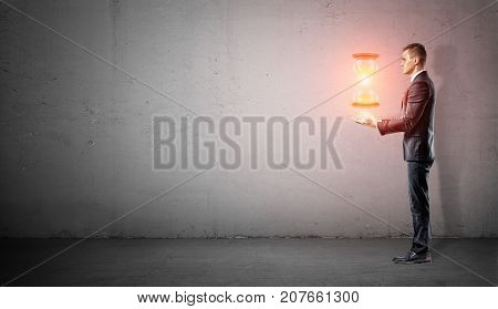 A businessman stands in side view on concrete background with his harms under a hovering glowing hourglass. Master of time. Keep time in check. Time control.