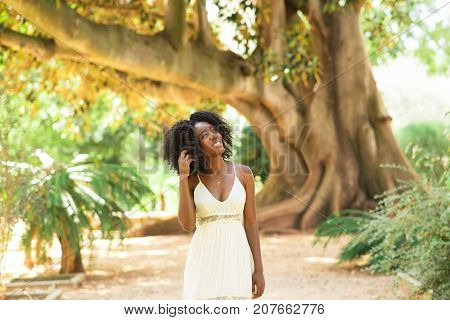 Portrait of smiling young attractive African American woman touching hair and walking in park with big tree in background