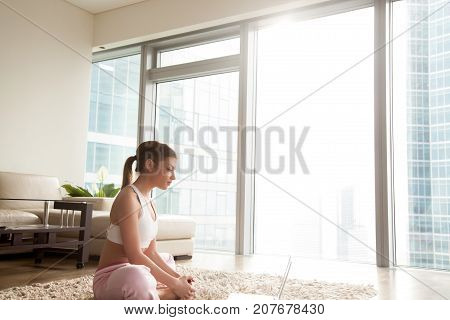 Young calm flexible woman sits in bound angle pose near laptop on floor at home interior, working out watching online yoga tutorial video, doing pilates exercise, baddhakonasana, butterfly stretch