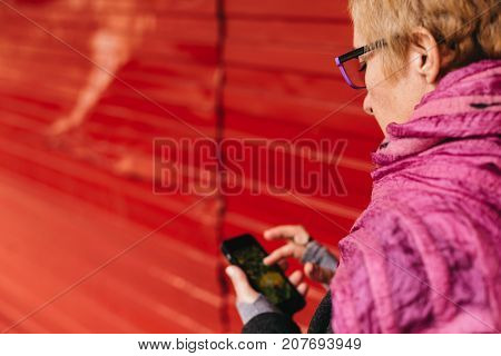Side view of adult blonde woman browsing smartphone standing at red fence wall.