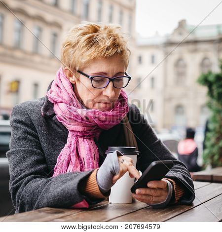 Adult blonde woman sitting with coffee and smartphone in outside cafe on street.