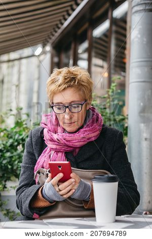 Smiling adult woman browsing smartphone and sitting with paper cup in outside cafe.