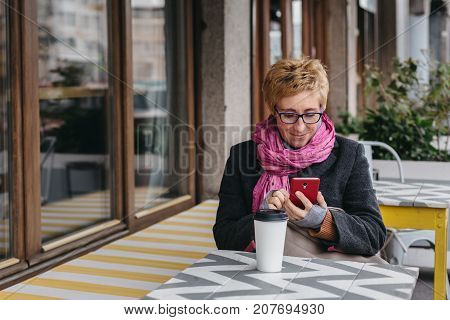 Adult blonde woman browsing smartphone and having drink from paper cup in outside cafe.