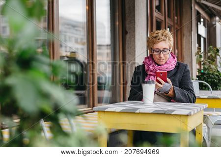Adult woman sitting at table in outside cafe and using smartphone while having coffee.