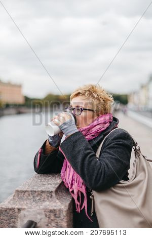 Side view of adult blond woman drinking coffee while standing at water on esplanade.