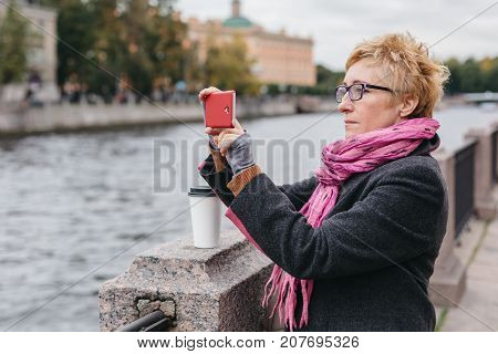 Adult woman standing on waterfront and taking shots with smartphone.