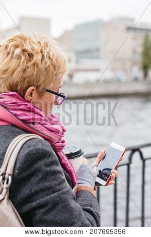 Adult woman in glasses standing with smartphone and coffee on waterfront.