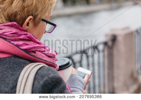 Adult woman in glasses standing with smartphone and coffee on waterfront.