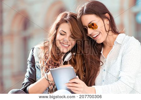 Portrait of two beautiful young girls sitting on bench looking at tablet having fun.