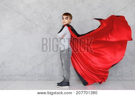 Boy profile portrait wearing Halloween vampire makeup and fluttering his red, black cloak, on a gray textured background. Cute kid wearing super hero costume with crossed hands, copy space available.