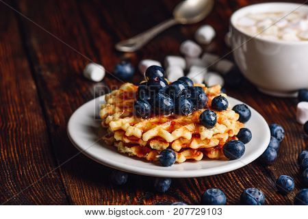 Homemade Waffles with Fresh Blueberry and Syrup on Plate. Cup of Coffee and Some Berries with Marshmallow on Backdrop.