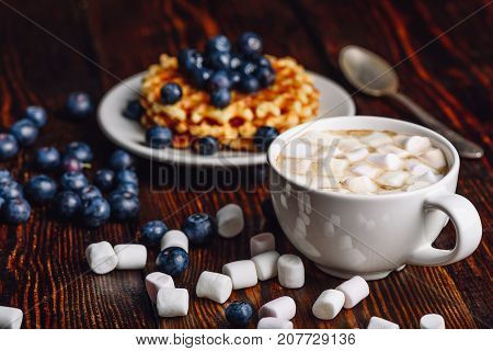 Cup of Hot Chocolate with Marshmallow with Belgian Waffles on Plate with Fresh Blueberry. Spoon and Some Berries and Marshmallows Scattered on Wooden Table.