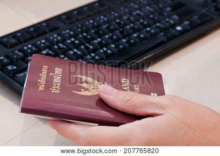 A hand holding Thailand passport besides computer keyboard.