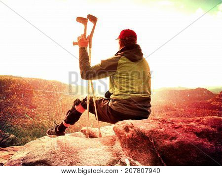Tired Tourist With Medicine Crutch  And Broken Leg Fixed In Immobilizer Resting On  Mountain Summit.