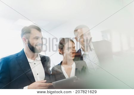 Two men and a woman in strict business suits are posing and smiling. They look at the glass wall hung with paper stickers. One of the men in the hands of a tablet