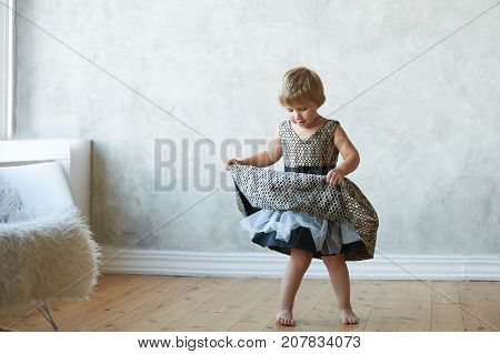 Indoor full lenght portrait adorable little girl with short hairstyle standing on floor barefooted and raising hemstitch of her new fancy dress dancing having happy expression on her cute face