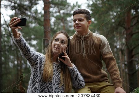 Happy siblings sister and brother taking selfie on mobile while hiking in woods. Beautiful young female holding smart phone taking self portrait posing in wild nature together with her boyfriend