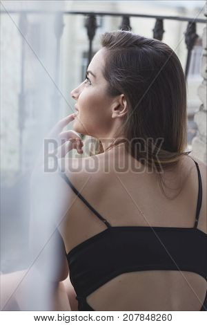 Vertical tender portrait of beautiful woman posing indoors through white curtain looking outside from indoor. Young brunette lady touching her softest skin feeling the moment enjoying fresh morning.