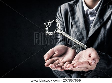 Cropped image of businessman in suit keeping big key in hands with dark wall on background.