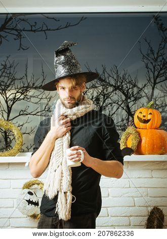 Halloween man holding cup at window with autumn trees. Macho wearing witch hat and scarf. Holiday symbols and decorations. Magic potion concept. Warm and homely atmosphere.