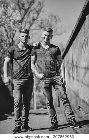 Twin brothers young handsome friendly blond men in jeans hug and smile outdoor on streetscape background