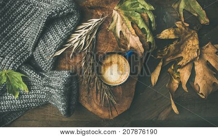 Autumn or Fall morning coffee concept. Flat-lay of arm knitted woolen grey sweater, wooden tray, mug of coffee and yellow fallen leaves over dark rustic wooden table background, top view