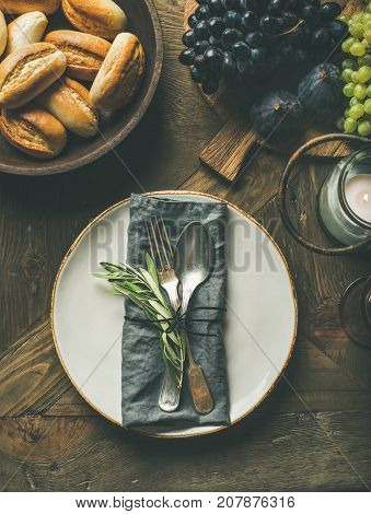 Fall holiday table decoration setting. Plate with linen napkin, fork and spoon, candle, fresh fruits on serving board, bread buns, olive tree branch over wooden background, top view