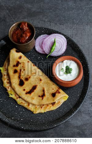 Aloo Paratha - Indian potato stuffed wheat bread served with Raita and mango pickle on dark moody background Top down view