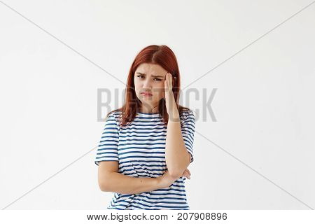 Frustrated young female in striped shirt holding head while suffering from bad headache or migraine because of stress and failure at work looking at camera with unhappy painful expression on her face