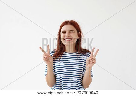 Body language. Horizontal studio shot of beautiful positive young female smiling broadly and making peace sign while posing in studio for picture. Teenage girl showing victory gesture with both hands