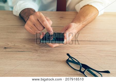 Smartphone in hands of successful businessman. Adult caucasian business person in white shirt with tucked rolled up sleeves using mobile phone at the desk in office.