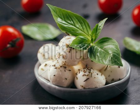Close up view of small mozzarella cheese marinated with herbs. Mozarella balls with fresh basil and cherry tomatoes on black concrete background. Low key.