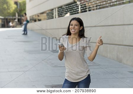 young beautiful and happy latin woman on urban street city background using internet on mobile phone texting and browsing social media in online communication concept giving thumb up
