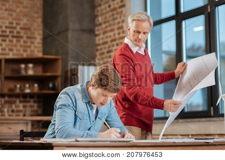 Quality-oriented. Pleasant senior man holding a blueprint and looking at the blueprint of his young colleague, comparing them