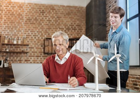 Pleasant ambience. Cheerful young man holding a blueprint and watching his senior colleague work on a laptop while holding a compass in his hand