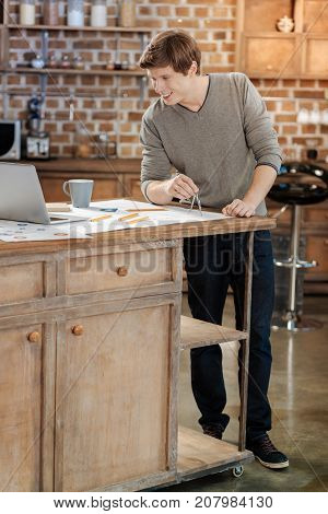 Learning new information. Handsome young man standing near the kitchen counter and drawing circles with a compass while watching a video tutorial on his laptop