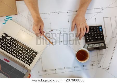 Work settings. The top view of male hands holding a pair of compasses and measuring distance on a blueprint while a laptop, a calculator and a cup of tea standing on it