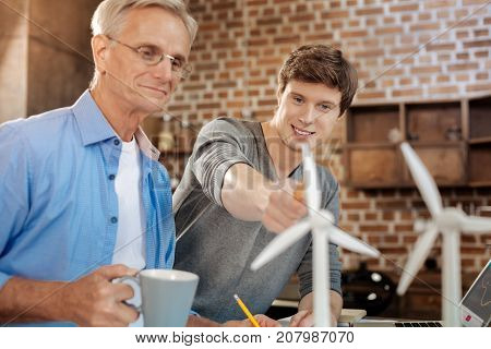 Smart remark. Pleasant young man pointing at a wind turbine model and making a remark about it while his mentor listening to it and smiling with approval