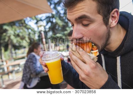 A Guy Is Greedily Eating His Cheesburger, His Eyes Are Closed. He Is Holding Fruit Juice At His Righ