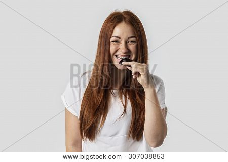 Young Red-haired Girl Isolated On Gray Background In White T-shirt. She Bites A Credit Card. The Con