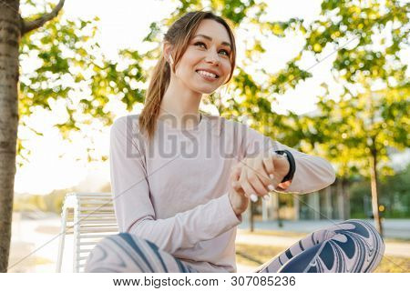Cheerful young fitness woman relaxing outdoos, sitting on a bench, using smart watch