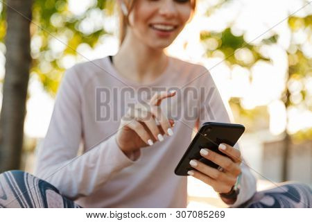Close up of a cheerful young fitness woman relaxing outdoos, sitting on a bench, using mobile phone