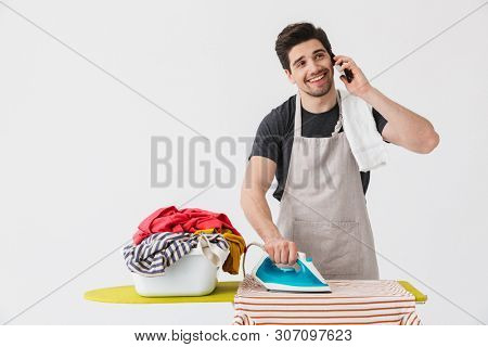 Image of a young happy houseman househusband with iron near clothes isolated over white wall background talking by mobile phone.