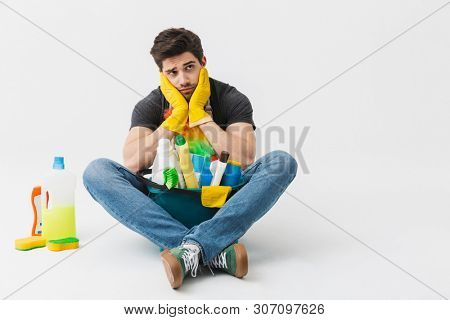 Image of a tired bored young houseman househusband with cleansers on floor isolated over white wall background.