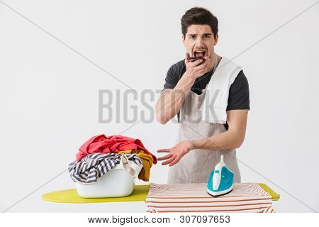Image of a young confused displeased houseman househusband with iron near clothes isolated over white wall background talking by mobile phone.